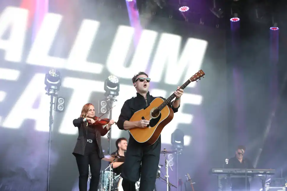 Callum Beattie plays acoustic guitar on stage with his band blurred behind him