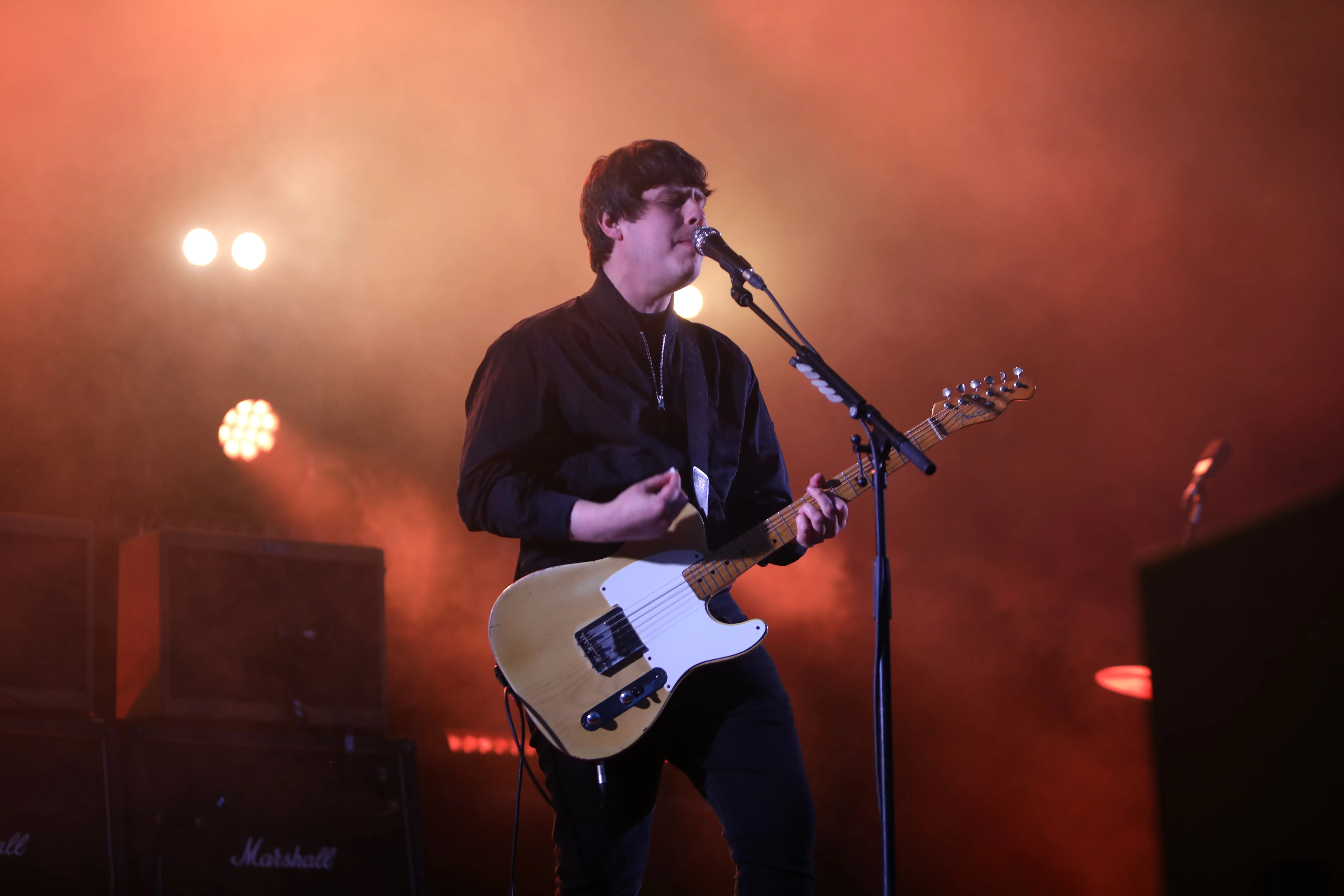 Jake Bugg strums his electric guitar and sings in front of Marshall amps and a dramatically lit stage